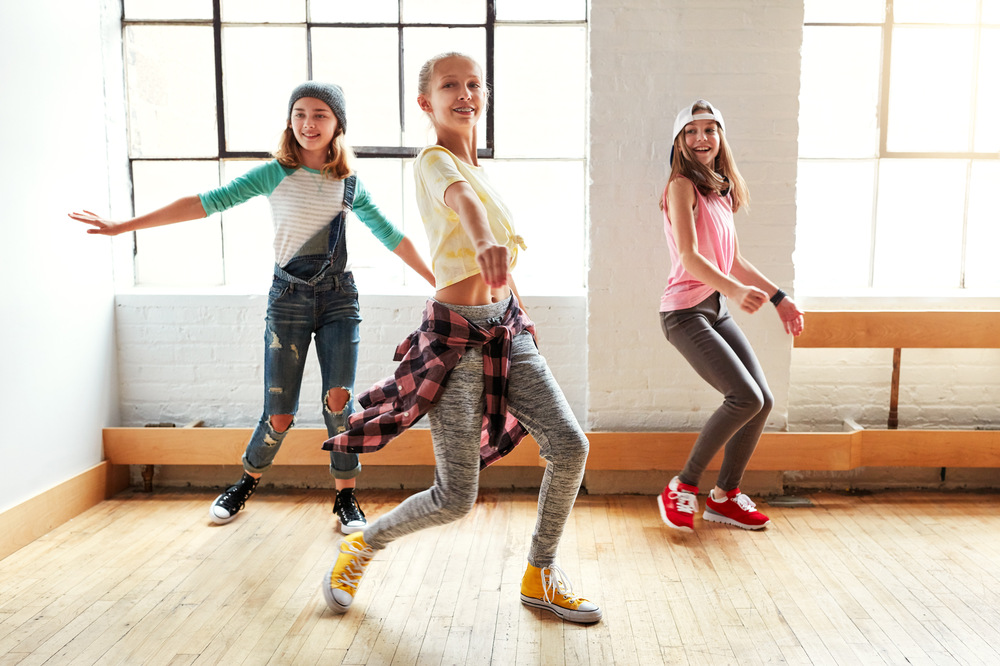 Tre unga personer dansar i ett ljust rum med stora fönster och trägolv.