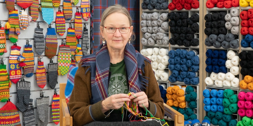 En kvinna sitter och stickar i en garnbutik omgiven av färgglada garnnystan och stickade vantar på väggen.