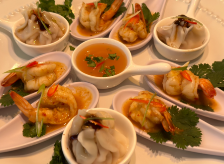 Små skedar med räkor, dumplings och koriander runt en skål med sås på ett vitt fat.
