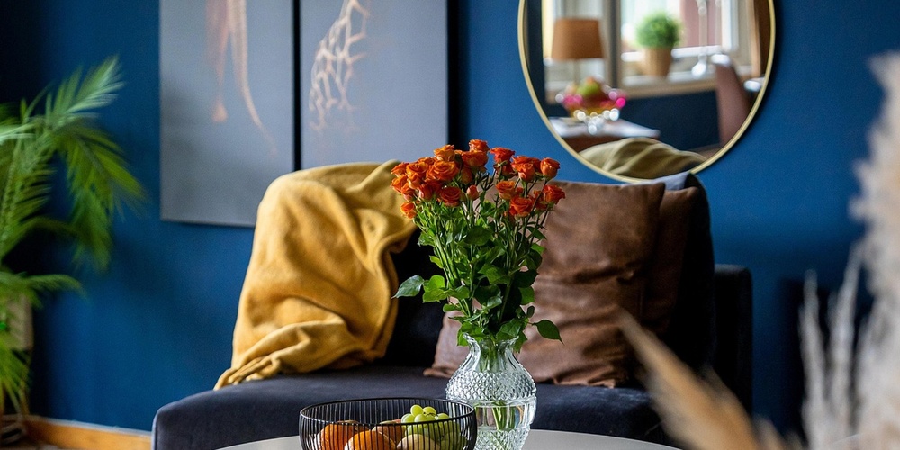 Stilfullt vardagsrum med blå väggar, rund spegel, soffor, blommor på bordet och djurtavlor på väggen.