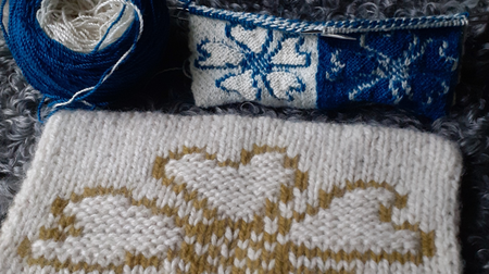 Två stickade provlappar med hjärt- och snöflingemönster i blått, vitt och beige garn.