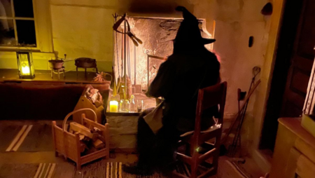 En person i svart häxhatt sitter vid en öppen spis i ett svagt upplyst rum med träinredning.