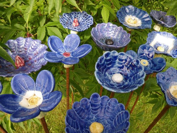 Blå keramikblommor på pinnar står i en trädgård med gröna blad i bakgrunden.