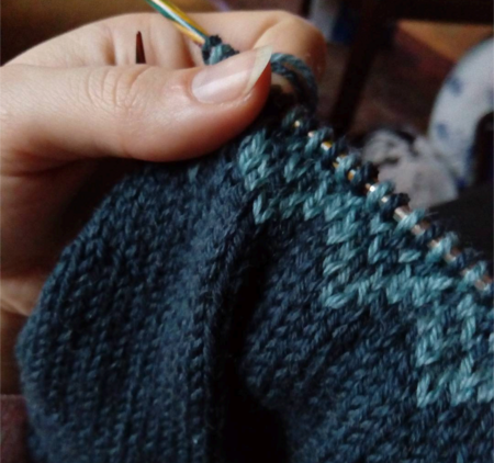En närbild på en hand som håller i en stickning. Mönstret är mörkblått och har en ljusblå kant högst upp.
