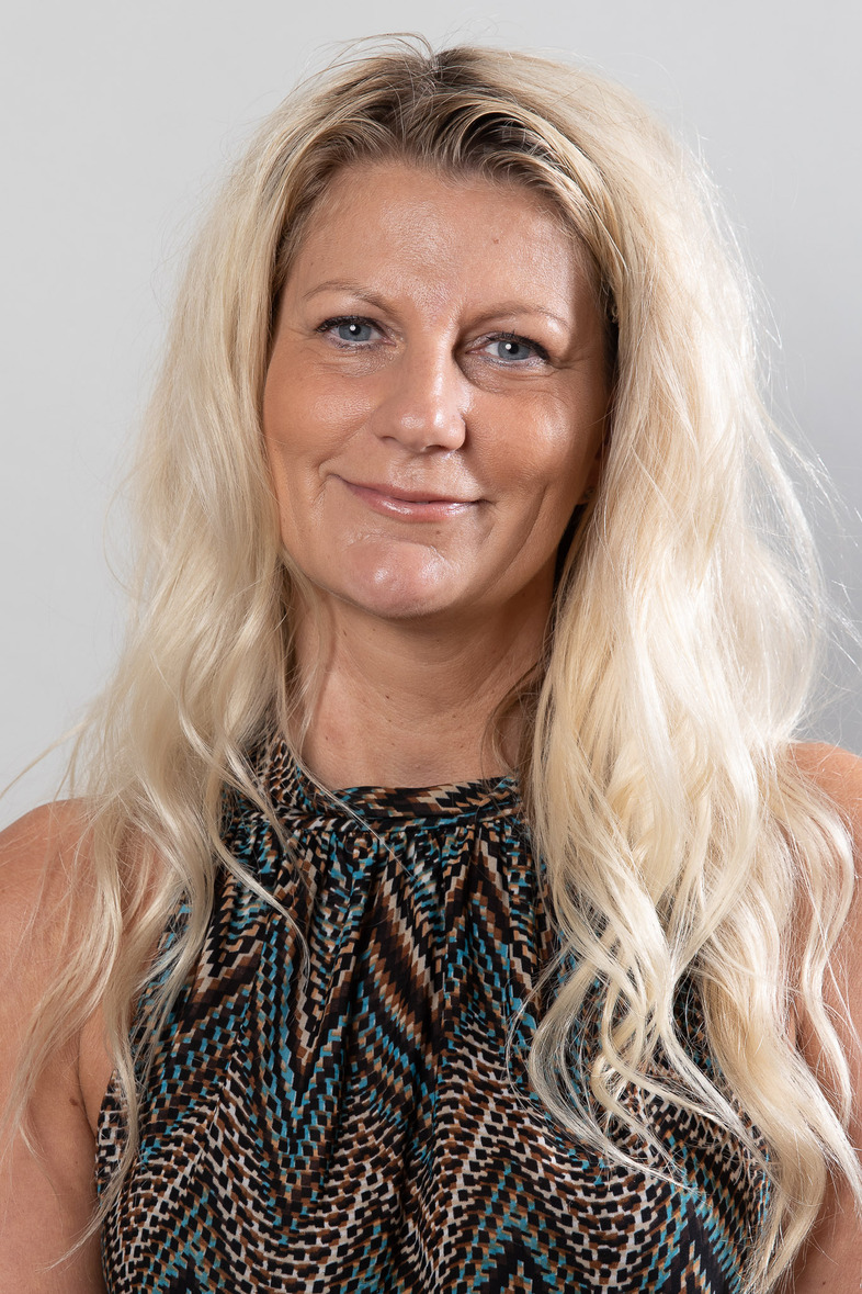 Kvinna med långt blont hår och mönstrad topp står framför en ljus bakgrund och ler mot kameran.
