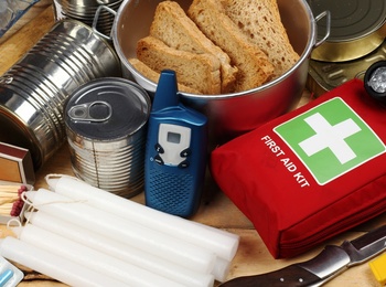 Nödutrustning med konserver, vattenflaskor, första hjälpen-kit, ficklampa, kniv, ljus och mediciner på ett träbord.