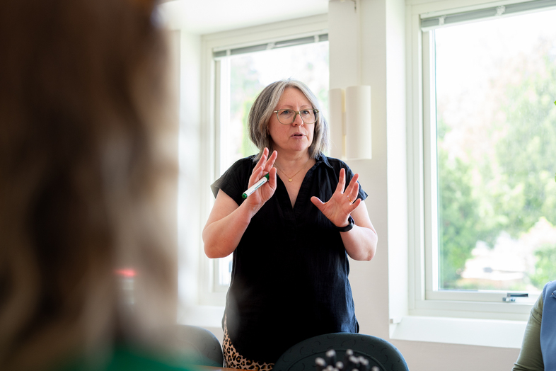 Kvinna med glasögon står och pratar framför ett fönster i ett ljust rum.