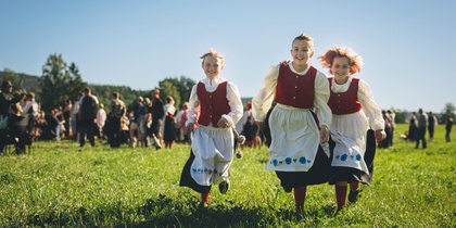 Tre barn i folkdräkt springer glada över en grön äng med en grupp människor i bakgrunden.