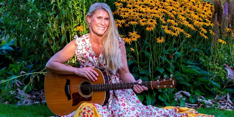 Kvinna i blommig klänning sitter på gräset och spelar gitarr framför gula blommor i en trädgård.