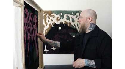 Person med tatueringar arbetar med tuftning framför en färgglad textilvägg.
