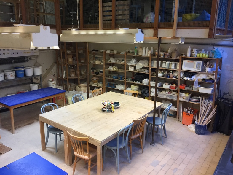 Ett keramikverkstad med stora arbetsbord, stolar och hyllor fyllda med material och verktyg.