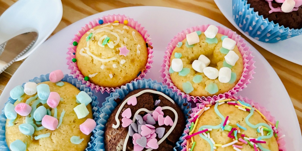 Flera muffins med färgglada strössel och minimarshmallows på vita fat på ett träbord.