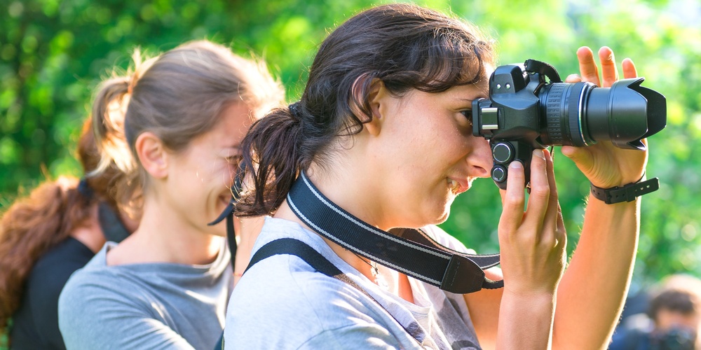 Två kvinnor fotograferar utomhus i grönskande miljö, med en tredje person i bakgrunden.