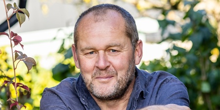 En person står i en trädgård med armarna vilande på ett trädgårdsredskap, omgiven av gröna växter.