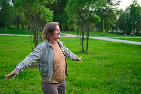 Person står med utsträckta armar i en grön park, ser glad ut och njuter av naturen.