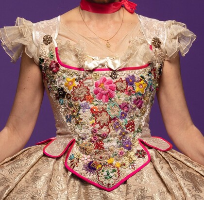 Närbild av en dekorerad, blommig korsettklänning med puffärmar och rosa detaljer mot lila bakgrund.