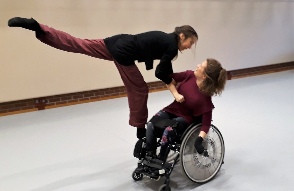 Två personer dansar tillsammans, en sitter i rullstol och den andra balanserar med ett ben i luften.