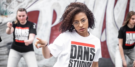 Tre unga personer dansar framför en graffitivägg, alla bär t-shirts med texten 'DANS STUDIO'.