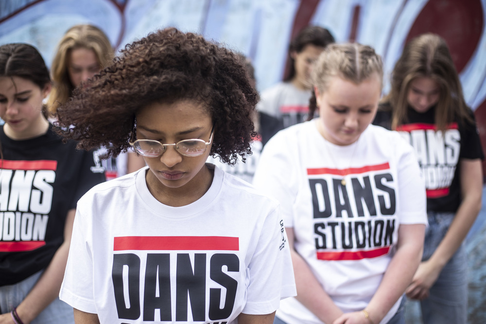 En grupp unga personer står med huvudet böjt, alla bär t-shirts med texten 'DANS STUDIO'.