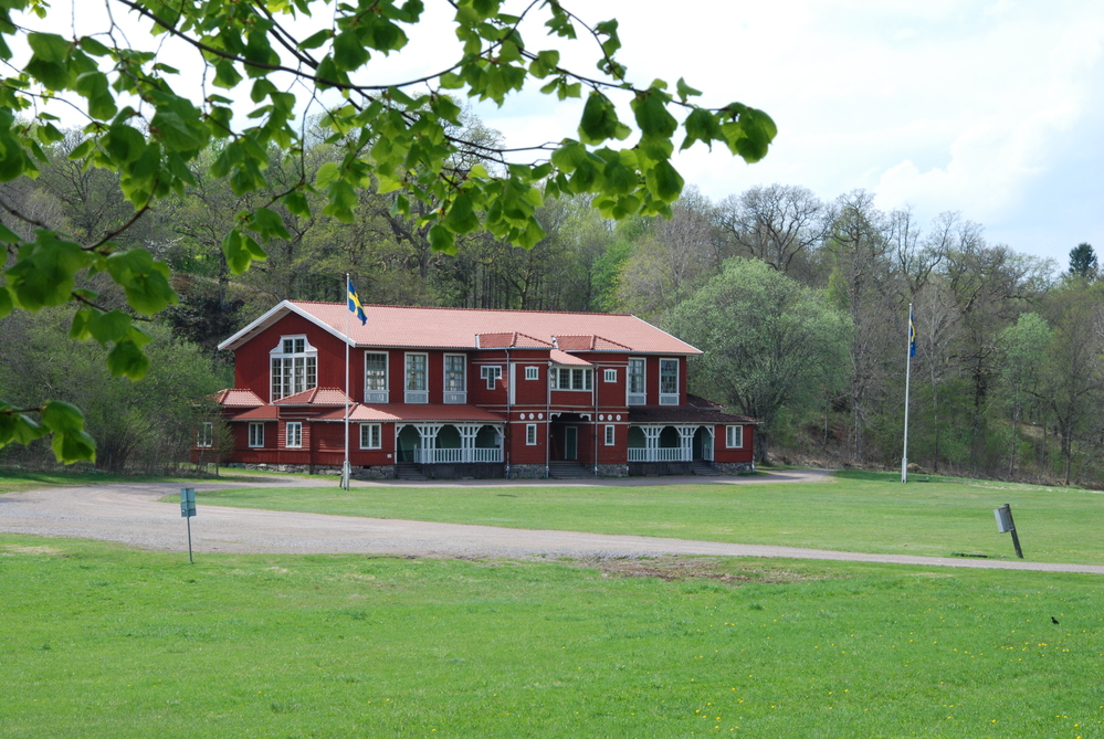 Röd trävilla med vita detaljer och svenska flaggor, omgiven av grön gräsmatta och träd.