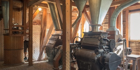 Interiör av en gammal kvarn med stora maskiner och träbjälkar, svagt ljusinsläpp från fönster.