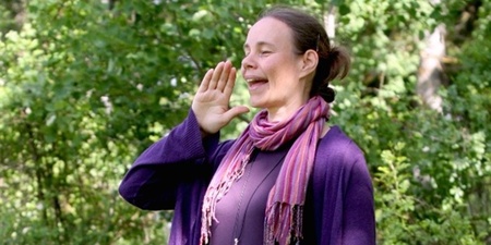 Kvinna i lila kläder och halsduk ropar eller sjunger utomhus bland gröna träd.