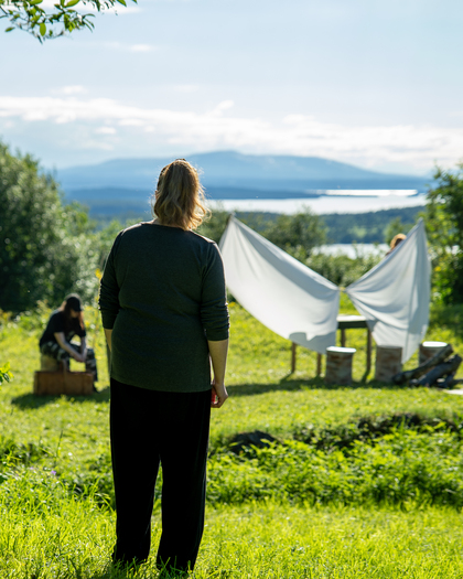 En person står på en gräsmatta och tittar mot två andra vid ett vitt tyg utomhus, med berg och sjö i bakgrunden.