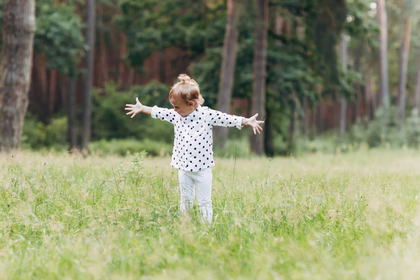 Ett litet barn med utsträckta armar står på en äng omgiven av högt gräs och träd.