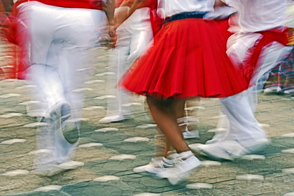 Personer dansar i rörelse, klädda i vita kläder och röd kjol, på en stenlagd gata.