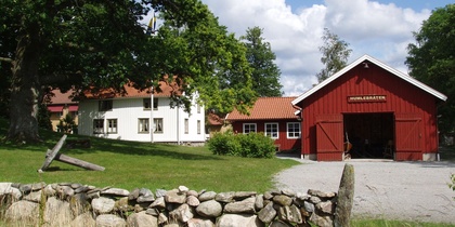 Gårdsmiljö med röd lada, vit byggnad, stengärdsgård och grönskande träd under blå himmel.