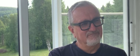 En äldre man med glasögon sitter vid ett bord inomhus med stora fönster och utsikt över grön natur.