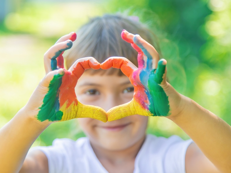 Ett barn håller upp händer målade i regnbågsfärger och formar ett hjärta framför ansiktet utomhus.