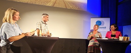 Fyra personer står vid två höga bord på en scen under en paneldiskussion med projektorbild i bakgrunden.