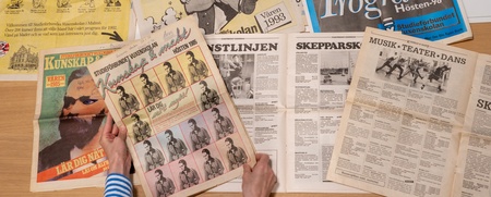 Flera gamla tidningar och programblad på svenska ligger utspridda på ett bord, en person bläddrar bland dem.