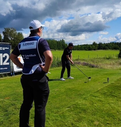 Två personer på en golfbana, en slår ut från tee medan den andra, med namnet Bjerhag på västen, tittar på.