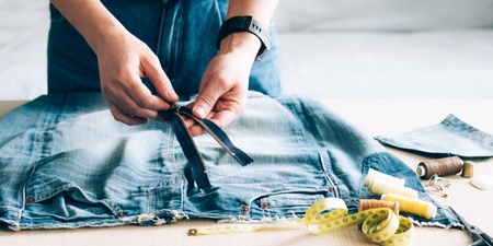 Person arbetar med att sy om ett par jeans, med dragkedja, måttband och sytråd på bordet.