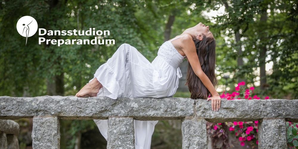 Kvinna i vit klänning poserar dansande på en stenmur utomhus med grönska och blommor i bakgrunden.