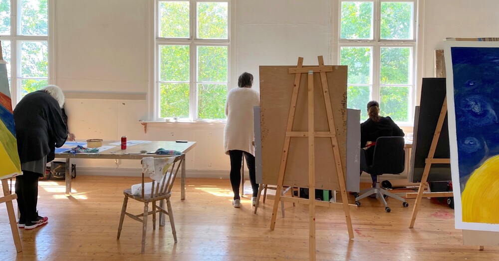 Tre personer arbetar med konst i en ljus ateljé med stora fönster och stafflier.