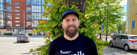 Man med skägg och keps står framför ett träd på en solig gata, bär svart t-shirt med texten 'musik'.