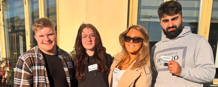 Fyra unga vuxna står utomhus i solskenet framför en gul byggnad, alla bär namnskyltar.