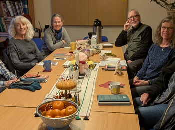 En grupp vuxna sitter runt ett konferensbord med fika, böcker och frukt i ett mötesrum.