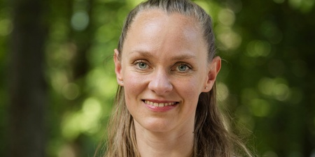 Person med långt blont hår och vit t-shirt står utomhus i en grönskande park.