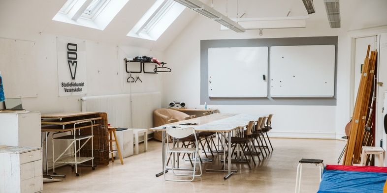 Ljust klassrum med bord, stolar, stafflier och vita tavlor på väggen. Inga människor syns i rummet.