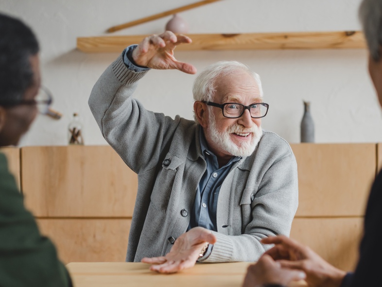 Tre äldre män sitter vid ett bord och samtalar, en av dem gestikulerar livligt med händerna.