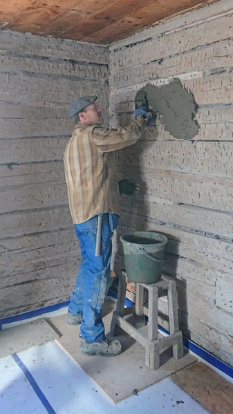 En person lagar en timmervägg med murbruk inomhus, står på en pall med en hink bredvid sig.