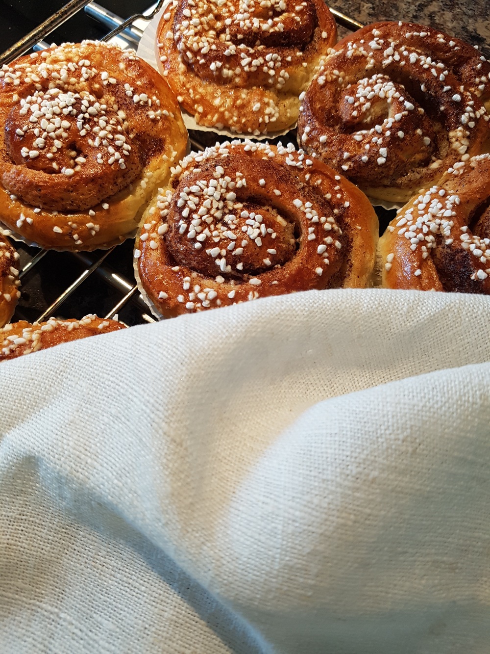 Nygräddade kanelbullar med pärlsocker ligger på ett galler, delvis täckta av en vit kökshandduk.
