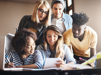 Fem unga vuxna studerar tillsammans vid ett bord, tittar på papper och ler.