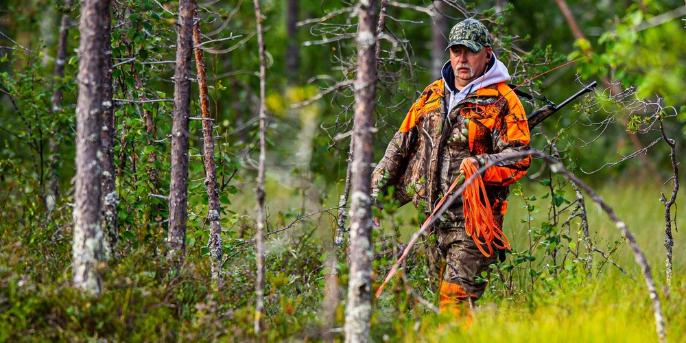 En jägare i orange och kamouflagekläder går med gevär genom en grön skog.