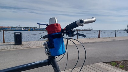 Närbild på ett cykelstyre med en blå och röd väska och en metallmugg, vid en kaj med vatten och fartyg i bakgrunden.