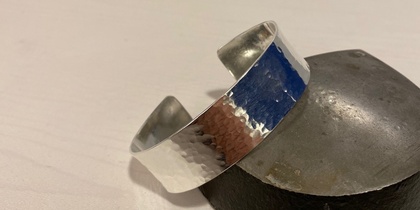 Ett silverfärgat, hamrat armband ligger lutat mot en metallklump på ett ljust underlag.